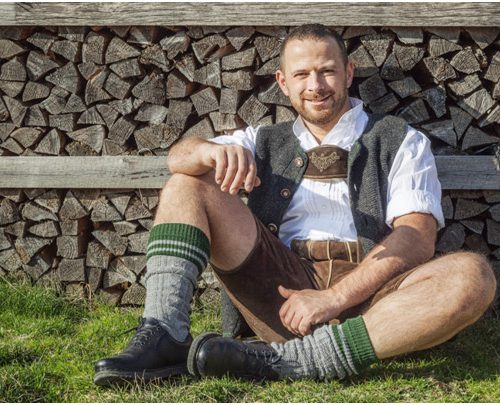 Mann in Lederhose bayrische Tracht, sitzend vor einem Holzstapel